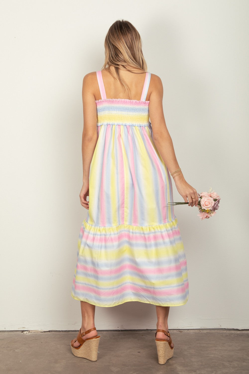 A woman in the VERY J Striped Woven Smocked Midi Cami Dress, featuring pastel stripes, holds a small bouquet of flowers. She pairs the dress with tan platform sandals that complete her chic summer look and stands against a plain white background.