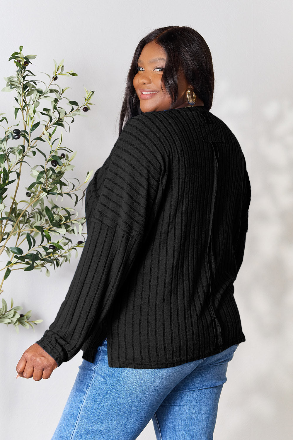 A person wearing the Basic Bae Full Size Ribbed Round Neck Slit Knit Top in blue and beige pants stands and smiles in front of a leafy plant.