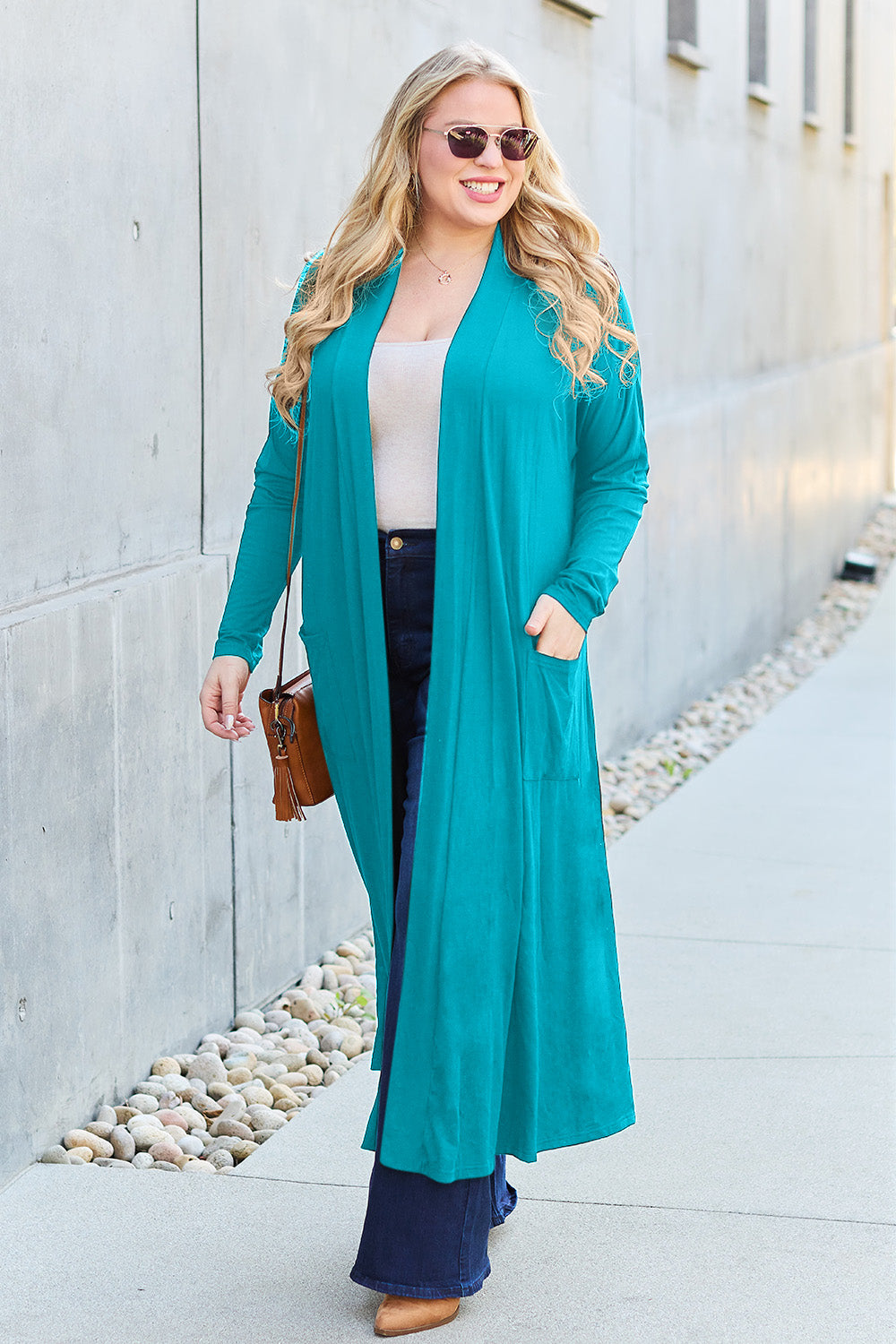 A woman stands against a concrete wall, wearing the Basic Bae Full Size Open Front Long Sleeve Cover Up made from rayon spandex, a white top, blue jeans in her perfect size, and brown shoes with her hands in her pockets. Machine wash cold for best results.