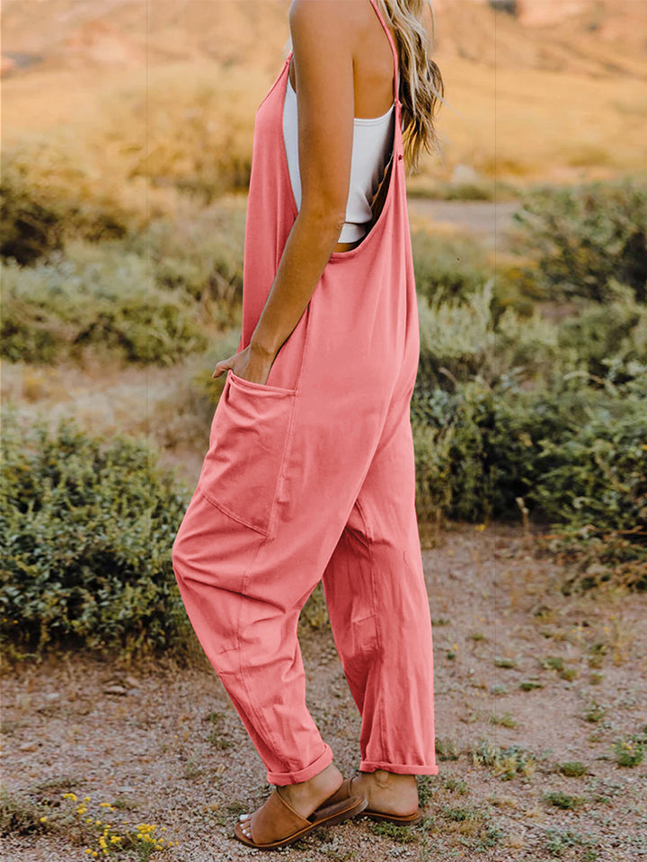 A woman wearing the Double Take Full Size Sleeveless V-Neck Pocketed Jumpsuit in coral and a beige cap is walking outdoors on a clear day, with a rocky landscape in the background.