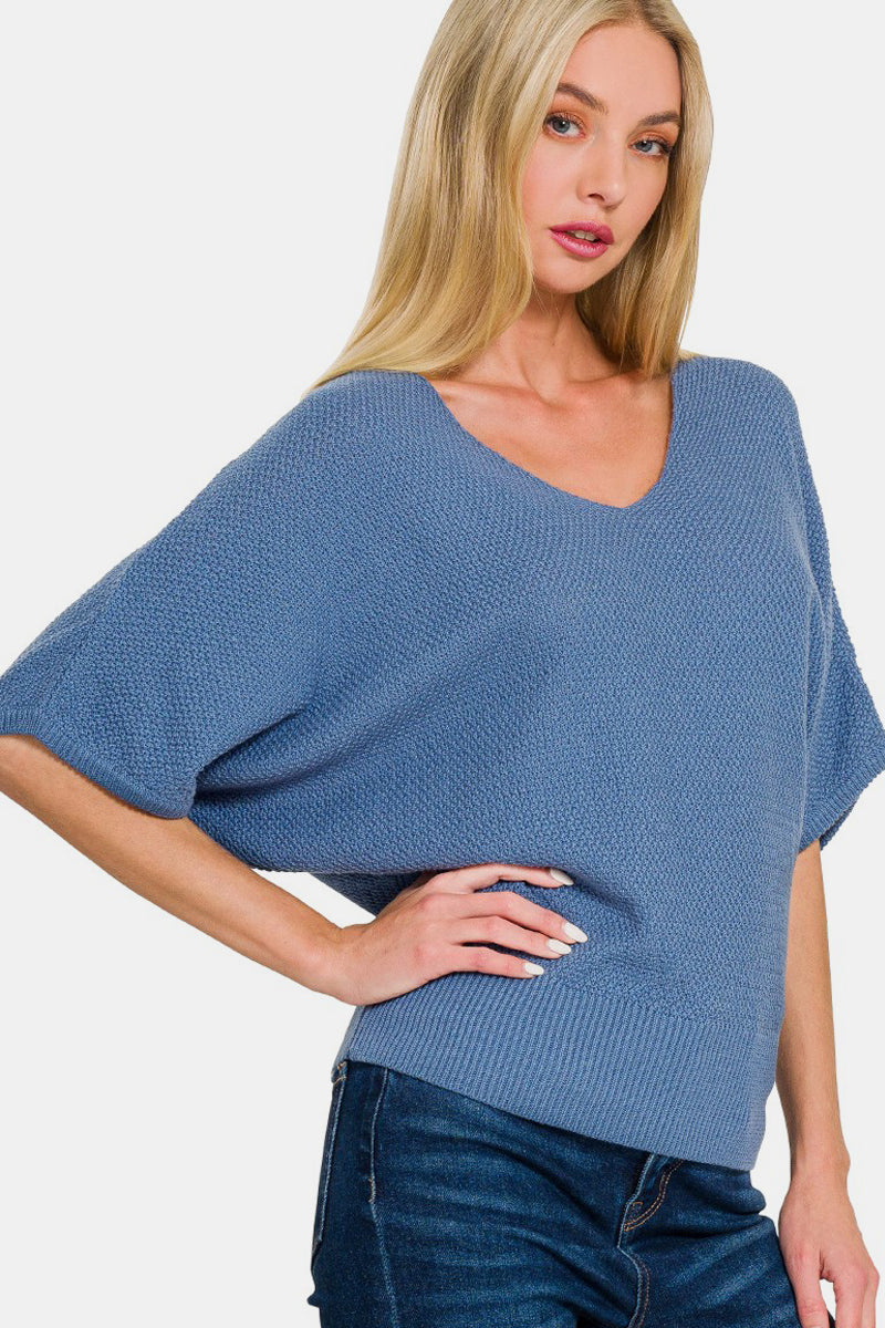 A woman with long blonde hair wears a loose-fitting Zenana V-Neck Short Sleeve Dolman Sweater in blue paired with dark blue jeans. She stands with one hand on her hip, looking towards the camera.