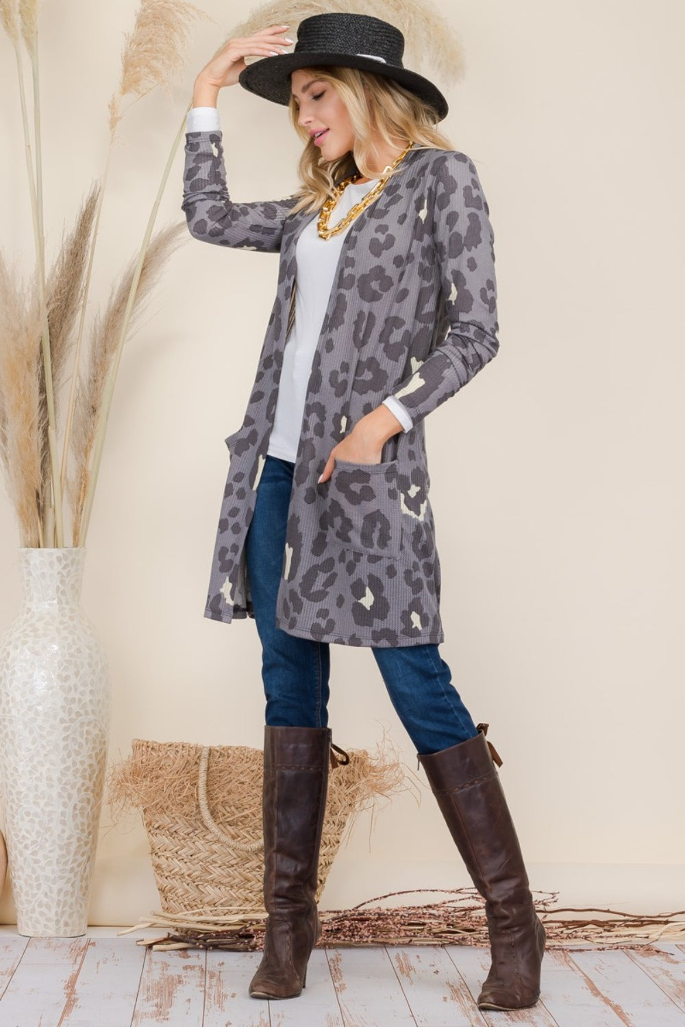 A person wearing a Celeste Full Size Leopard Open Front Contrast Cardigan over a white top and jeans, complemented by brown boots, accessorized with a black hat and necklace, stands next to a tall vase with decorative grasses.