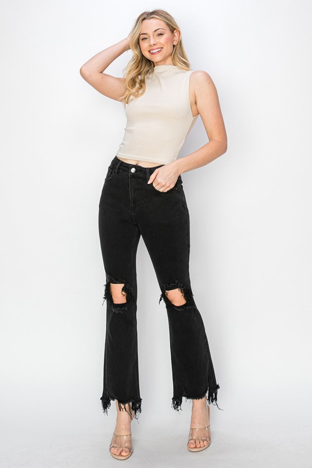 Rear view of a person wearing RISEN Full Size Distressed Raw Hem Jeans with Pockets in black, paired with clear-heeled sandals against a plain background.