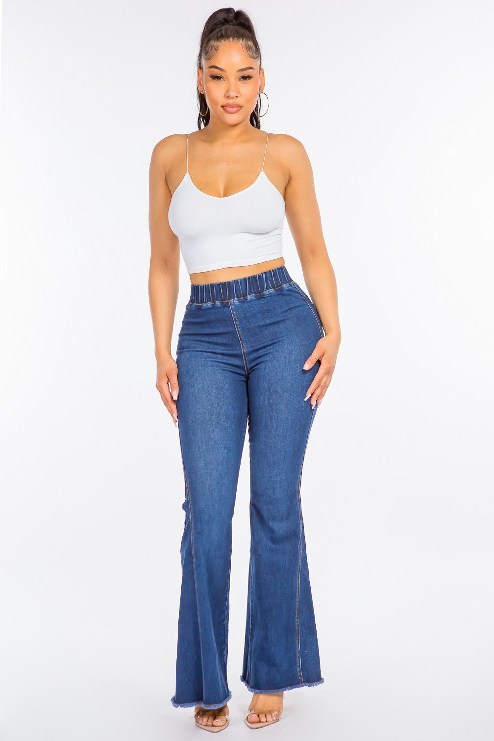 The image shows a person wearing American Bazi High Waist Curvy Flare Jeans made from stretchy denim, paired with nude open-toe heels. The jeans feature an elastic waistband and frayed hems, adding a touch of retro flair. The background is plain white.