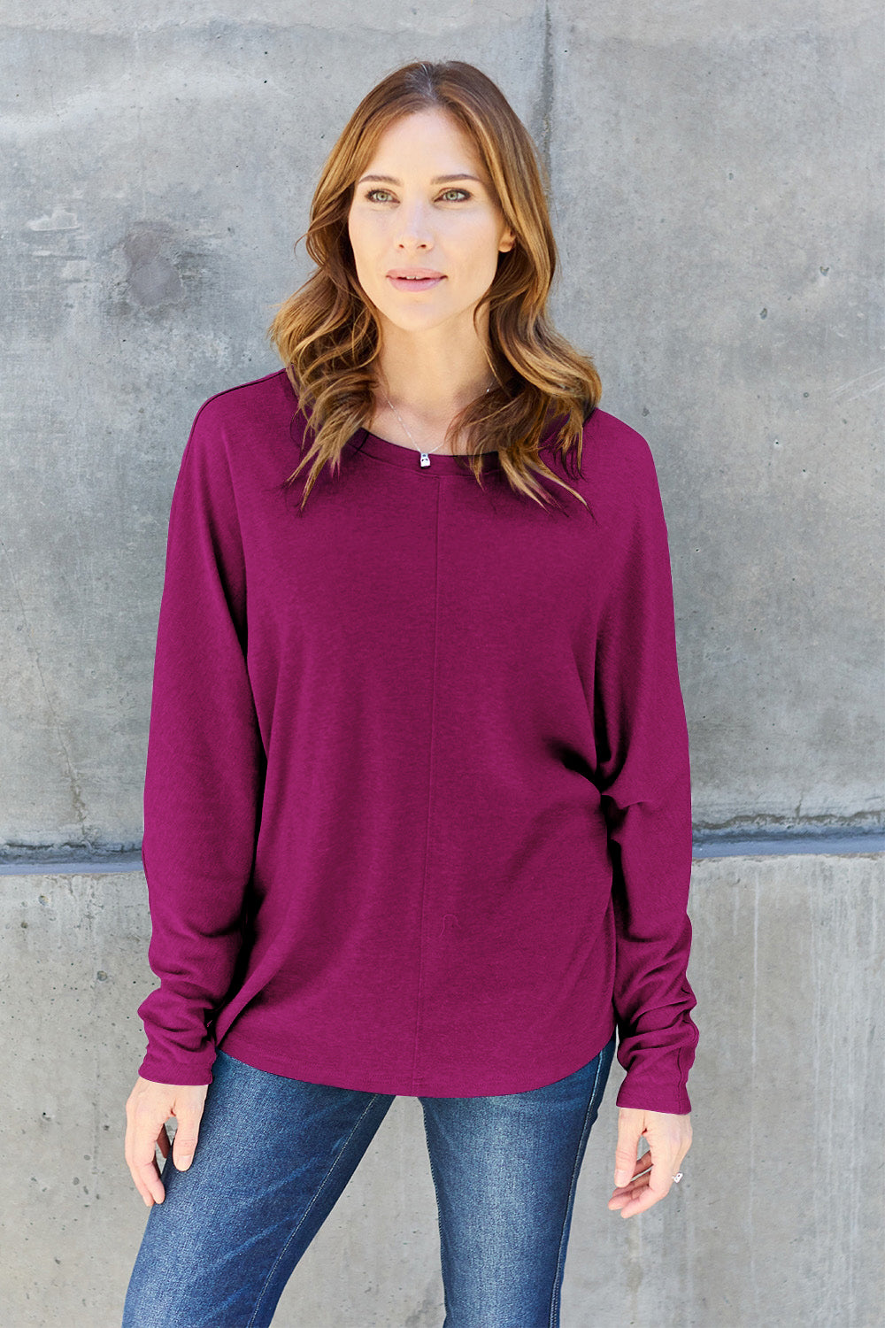 A person wearing a Double Take Full Size Round Neck Long Sleeve T-Shirt in green, blue distressed jeans with a slightly stretchy fit, and a crossbody bag, holding a beverage in a plastic cup.