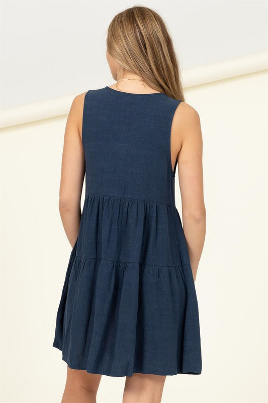 Wearing the Good Girl Sleeveless Tiered Mini Dress, a woman stands against a light background, gracefully holding the hem of her dark blue skirt with one hand.