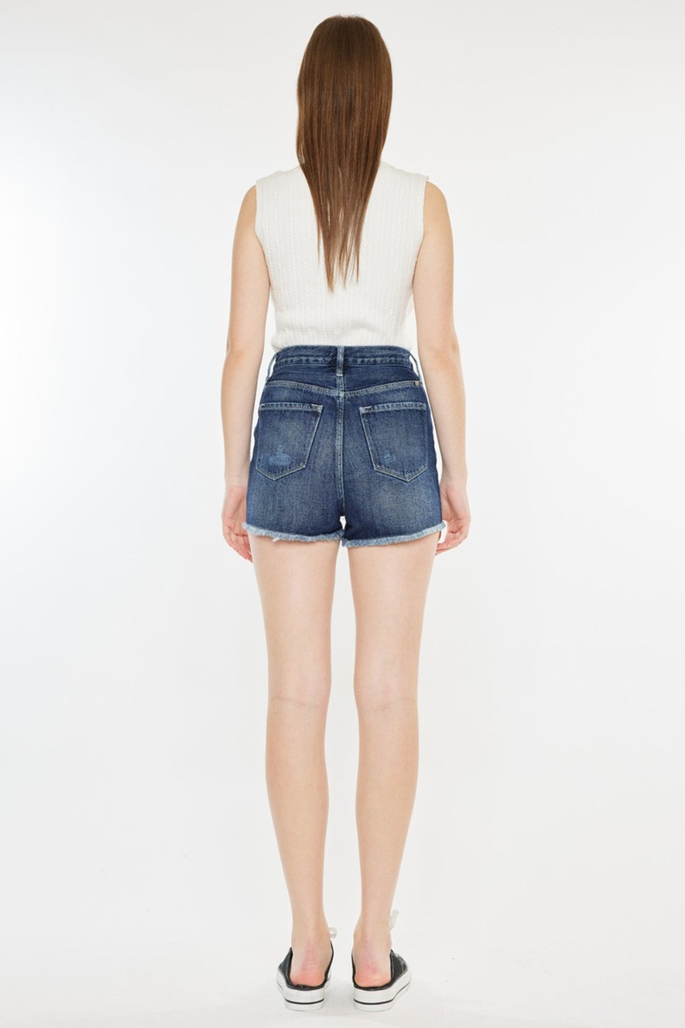 A woman stands against a plain background wearing a sleeveless white top, Kancan Raw Hem Button Fly Denim Shorts, and black slip-on sneakers.