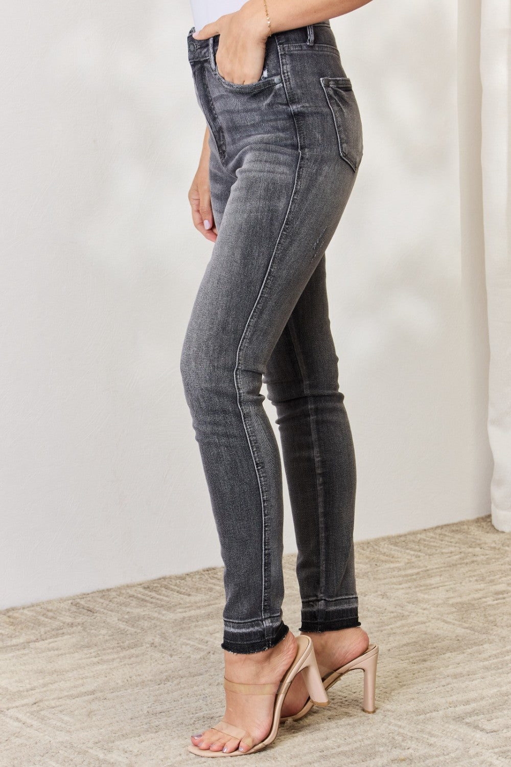 A person wearing the Judy Blue Full Size High Waist Tummy Control Release Hem Skinny Jeans paired with nude heels stands against a light background.