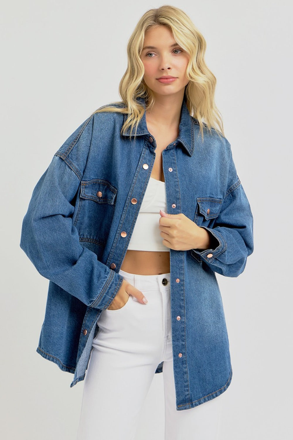 A person with long blonde hair, wearing the RISEN Full Size Snap Down Long Sleeve Denim Shacket over a white crop top and white pants, stands against a plain background.