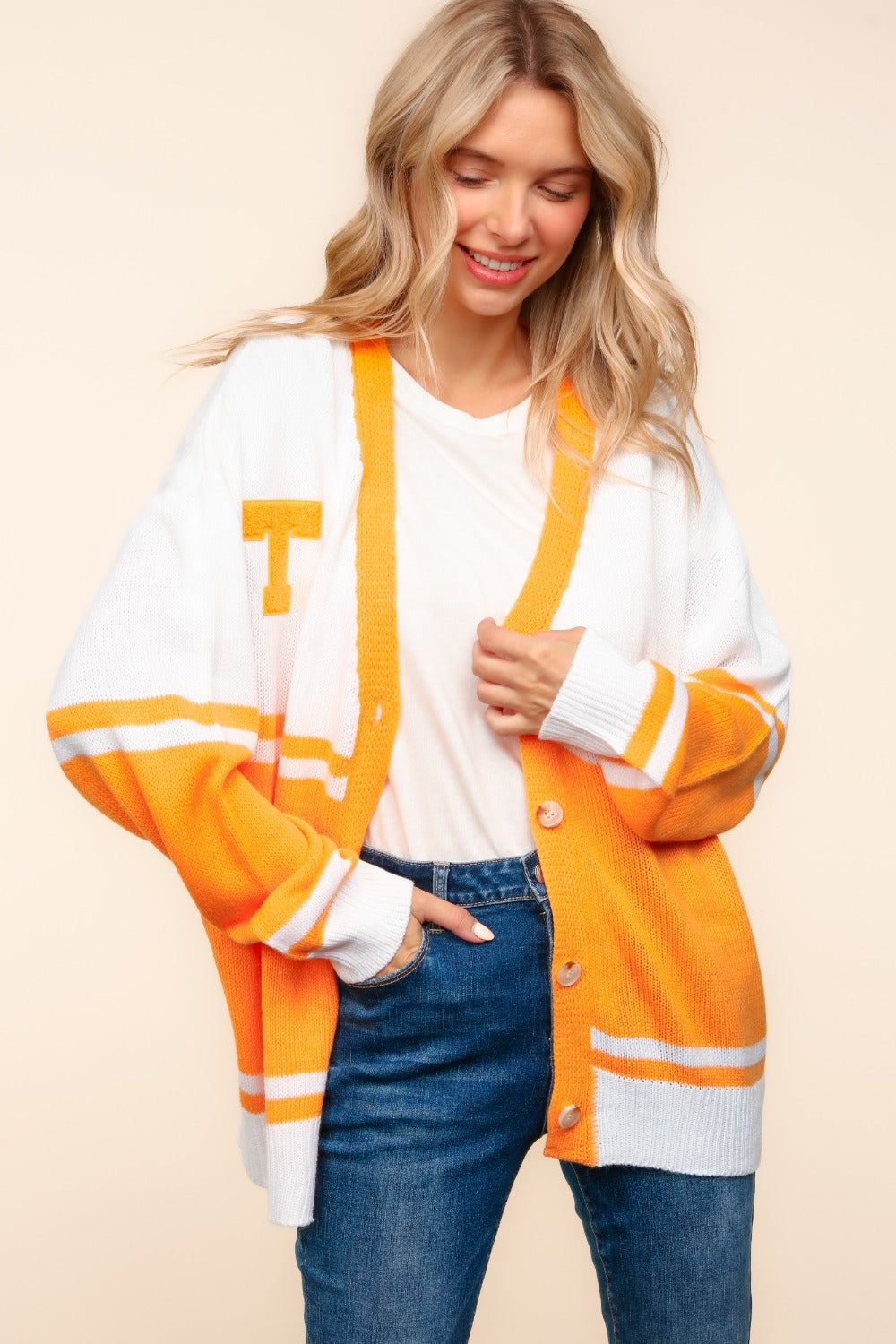 A person wearing the Haptics V Neck Button Down Letter Patch Cardigan, oversized with white and orange stripes and a yellow "T" patch, paired with blue jeans, smiles while lifting their hands to their head.