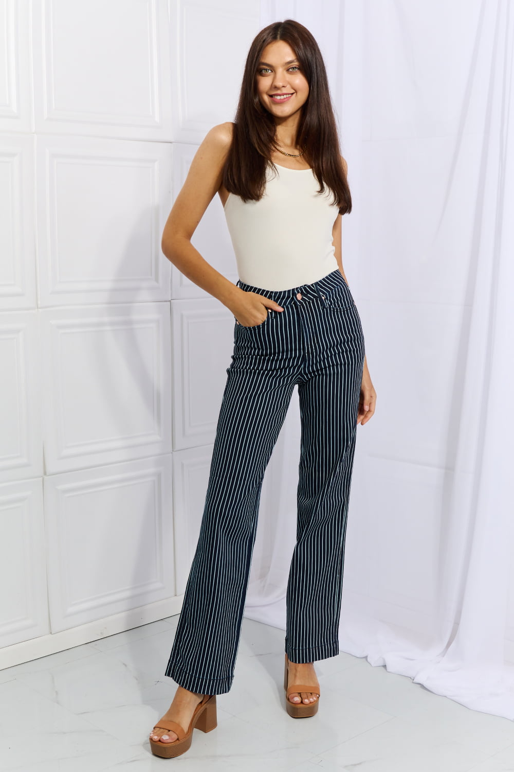 A person wearing Judy Blue Cassidy Full Size High Waisted Tummy Control Striped Straight Jeans, a white top, and tan platform sandals stands with one hand on their hip.