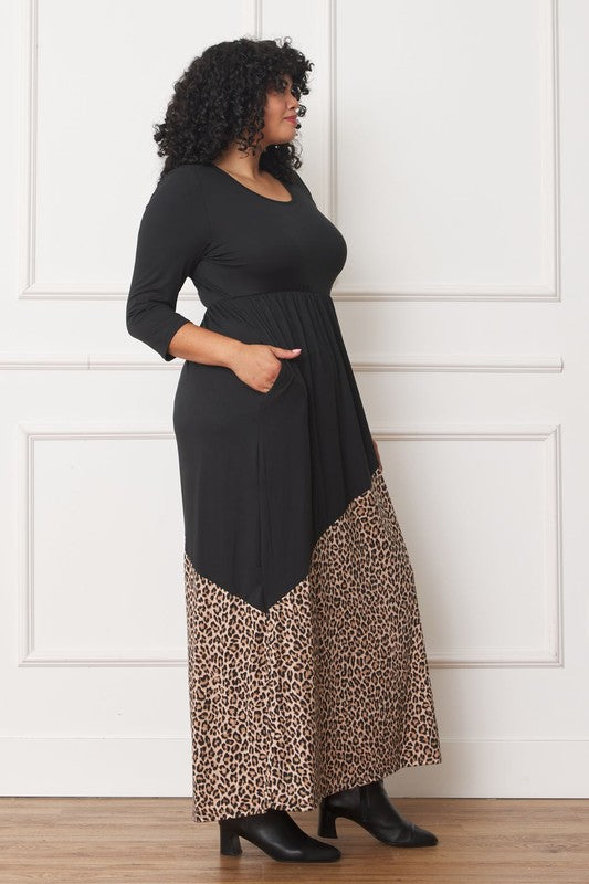 A woman with curly hair is wearing the Asymmetrical Leopard Accent Maxi Dress, featuring a long black design with a floral patterned section at the bottom. She stands against a white paneled wall, her hands tucked in her dress pockets, showcasing its elegant design.