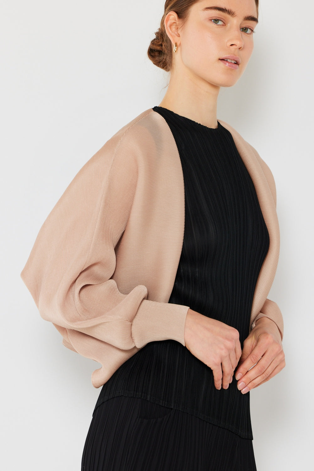 A person stands wearing the stylish Marina West Swim Rib Pleated Puff Sleeve Bolero Cardigan in beige, featuring draped sleeves, paired with a trendy black pleated top.