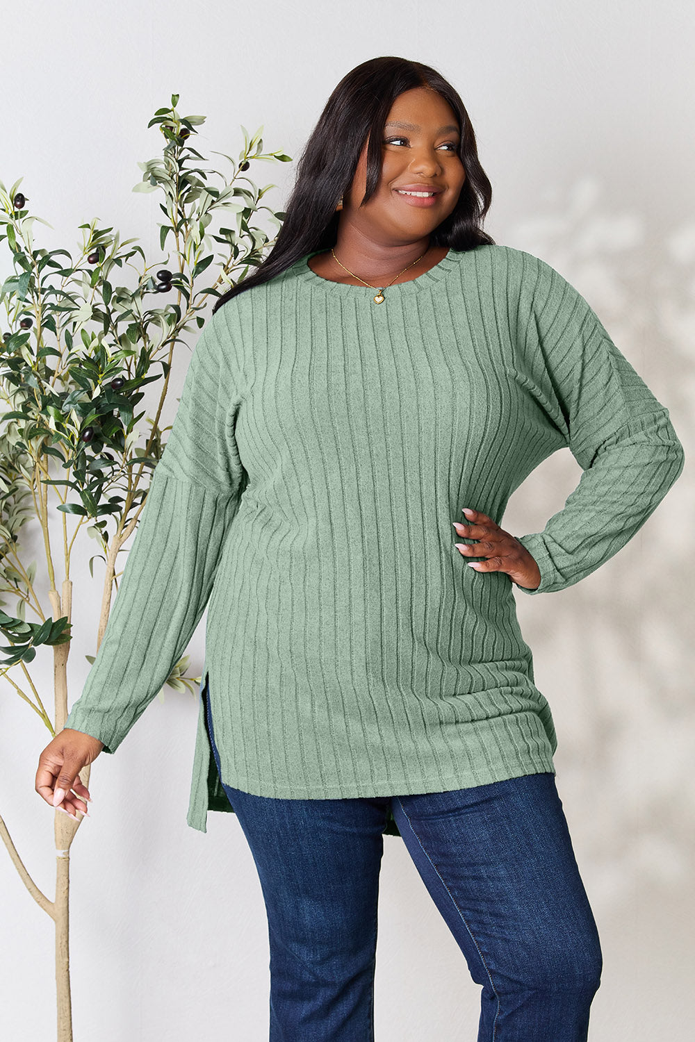 A person with shoulder-length hair smiles while wearing a beige ribbed sweater called the Basic Bae Full Size Ribbed Round Neck Long Sleeve Slit Top, along with matching beige pants, showcasing a basic style. They stand next to a green plant.