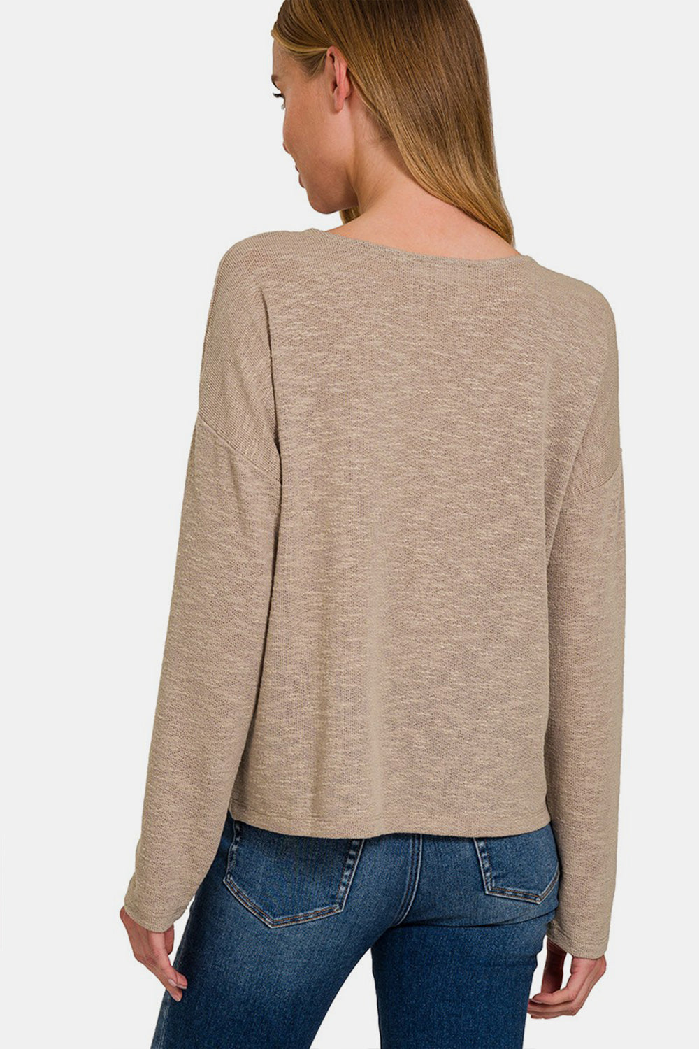 A woman with long blonde hair is wearing a beige Zenana Dropped Shoulder Long Sleeve T-Shirt and blue jeans, standing against a plain white background.