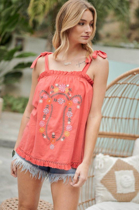 A person wearing an Embroidered Sleeveless Ruffle Tank Top and denim shorts stands outdoors near a wicker chair, adjusting their hair.