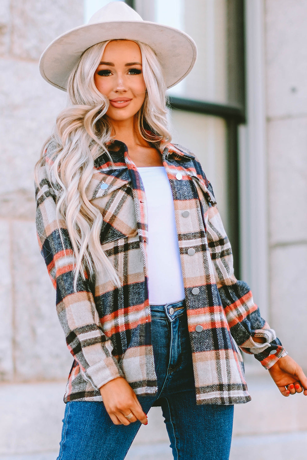 A person wearing a Brown Plaid Button Up Long Sleeve Flannel Shacket layered over a white top with ripped jeans and holding a brown handbag stands against a textured gray wall.