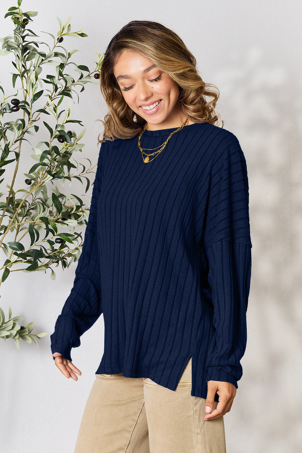 A person wearing the Basic Bae Full Size Ribbed Round Neck Slit Knit Top in blue and beige pants stands and smiles in front of a leafy plant.