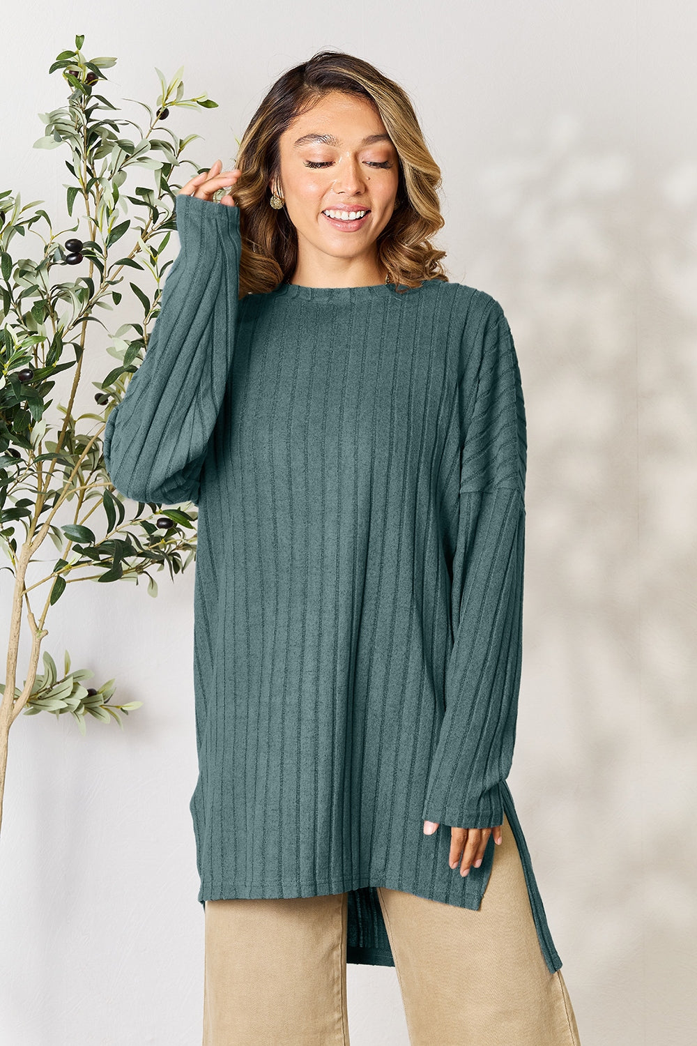 A person with shoulder-length hair smiles while wearing a beige ribbed sweater called the Basic Bae Full Size Ribbed Round Neck Long Sleeve Slit Top, along with matching beige pants, showcasing a basic style. They stand next to a green plant.