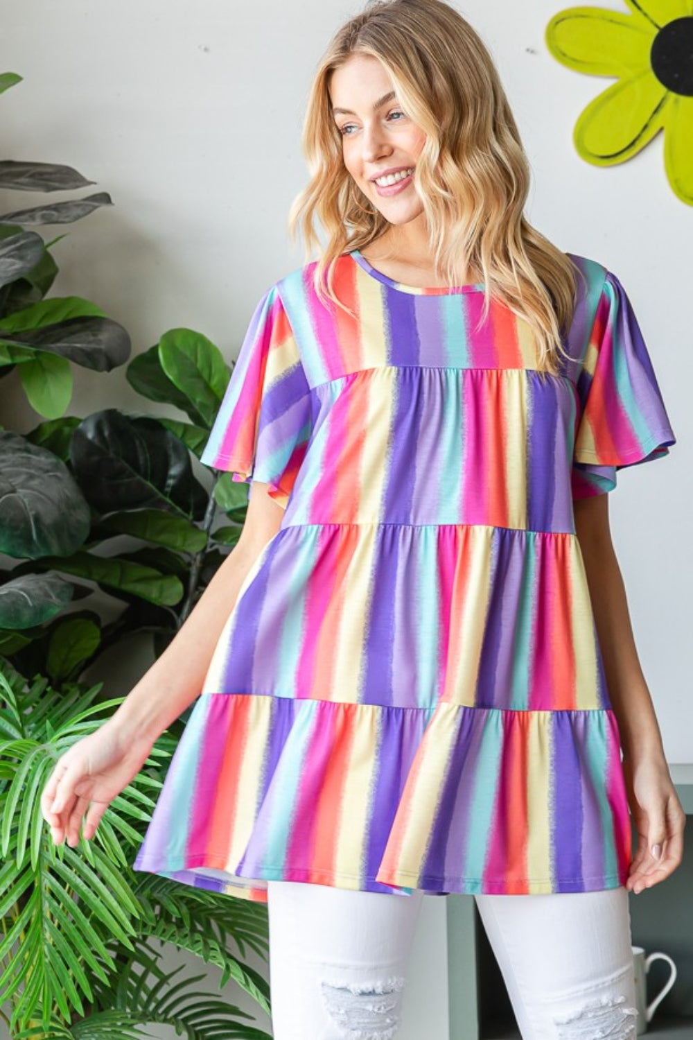 A person wearing the Heimish Full Size Short Sleeve Striped Tiered Top, featuring a colorful, classic striped pattern and flared sleeves, stands by a window. They are smiling, surrounded by plants in the background.