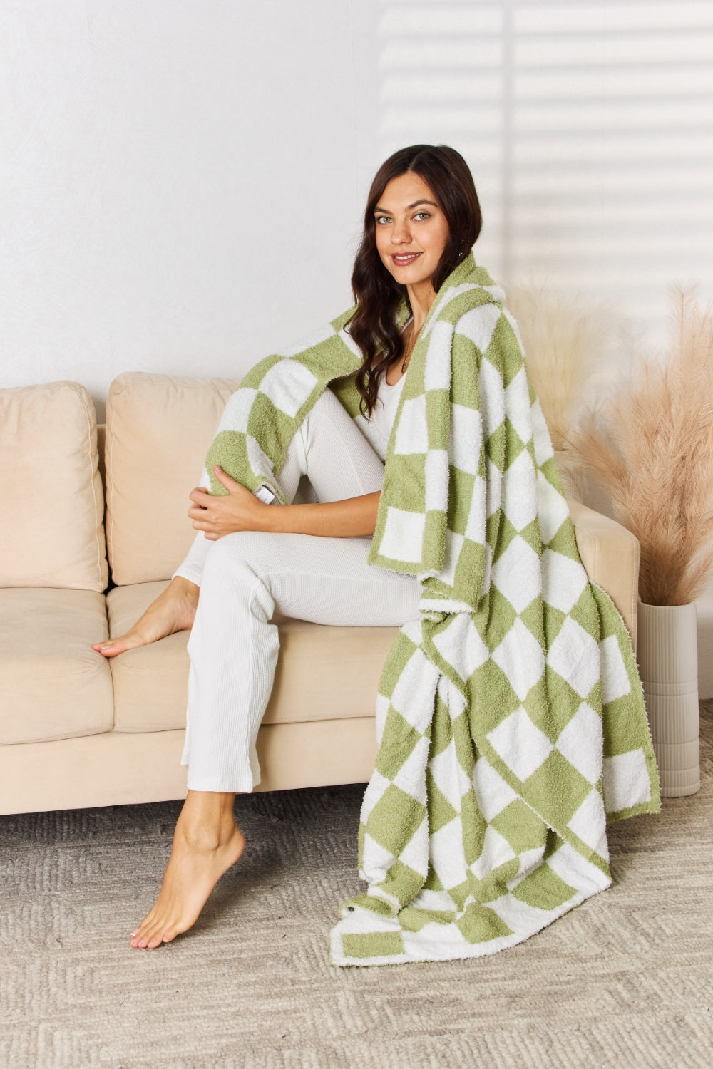 A person stands holding a large Cuddley Checkered Decorative Throw Blanket, smiling, in a room with a sofa and a potted plant. The blanket features a black and white checkerboard pattern, is made from 100% polyester, and imported for quality.