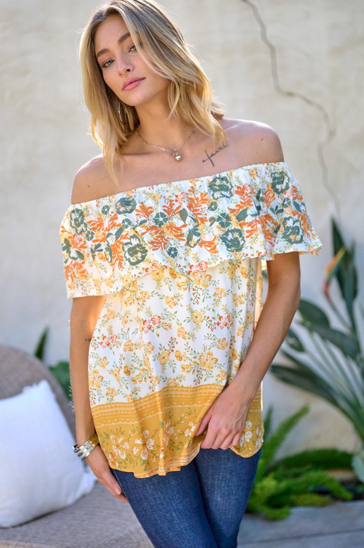 A woman with blonde hair is smiling while standing outdoors, wearing the Printed Off Shoulder Smocked Top paired with jeans.