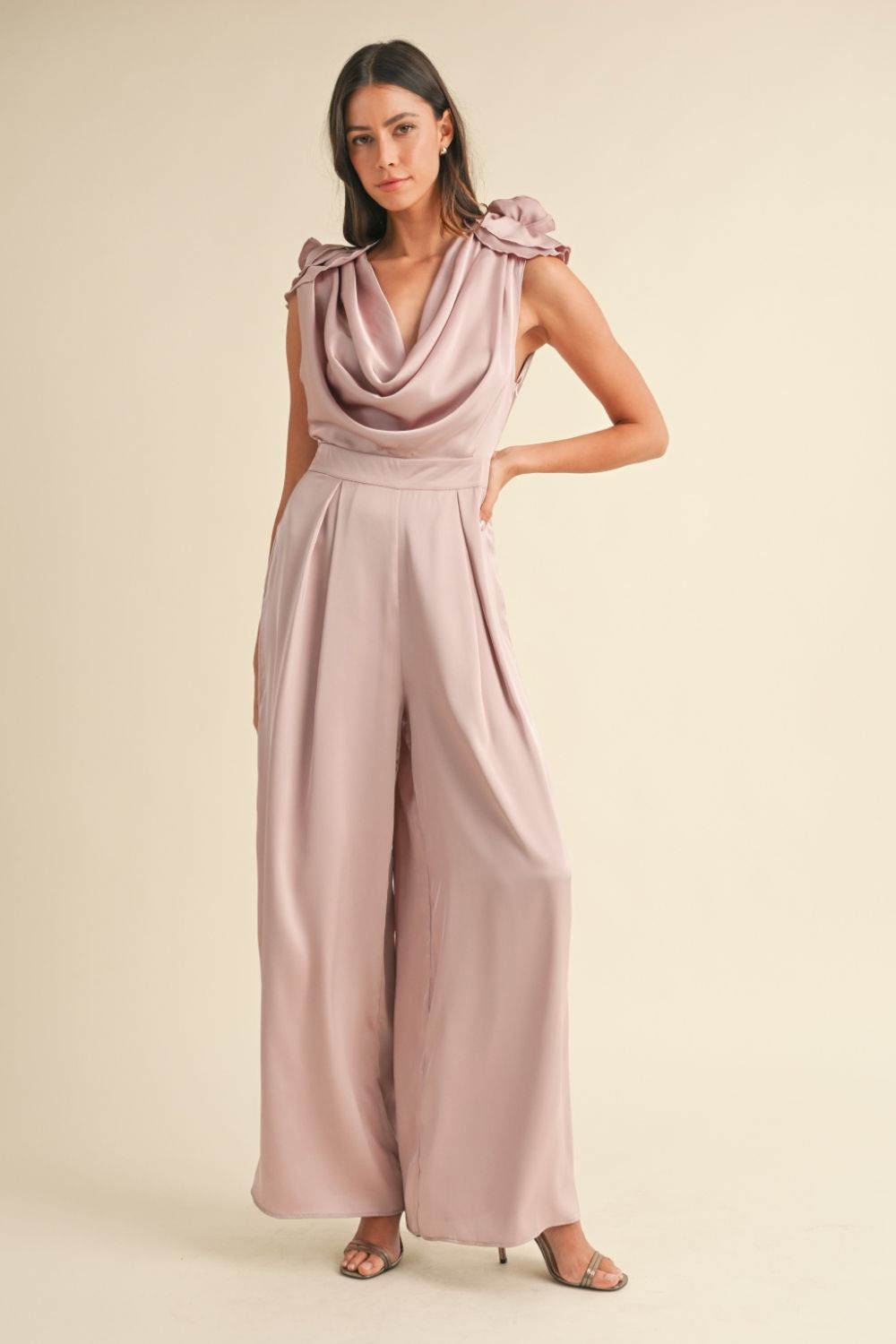 A woman stands against a plain background wearing the MABLE 3D Floral Applique Deep Cowl Neck Jumpsuit, which features an open back and wide-leg pants, paired with open-toed heels.