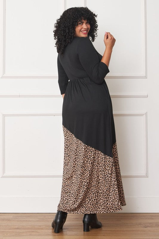 A woman with curly hair is wearing the Asymmetrical Leopard Accent Maxi Dress, featuring a long black design with a floral patterned section at the bottom. She stands against a white paneled wall, her hands tucked in her dress pockets, showcasing its elegant design.