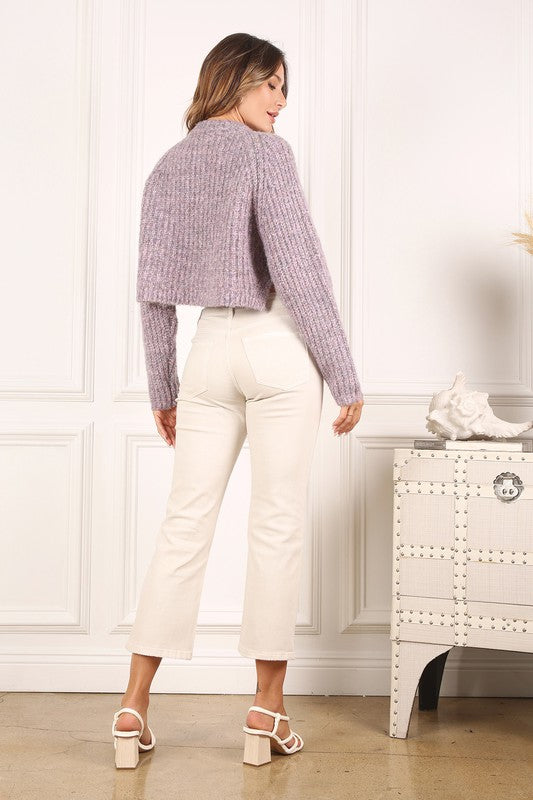 A person is wearing the Melange multicolor sweater top, characterized by textured pink fabric and featuring raglan sleeves along with large buttons, paired with light blue jeans, standing against a plain background.