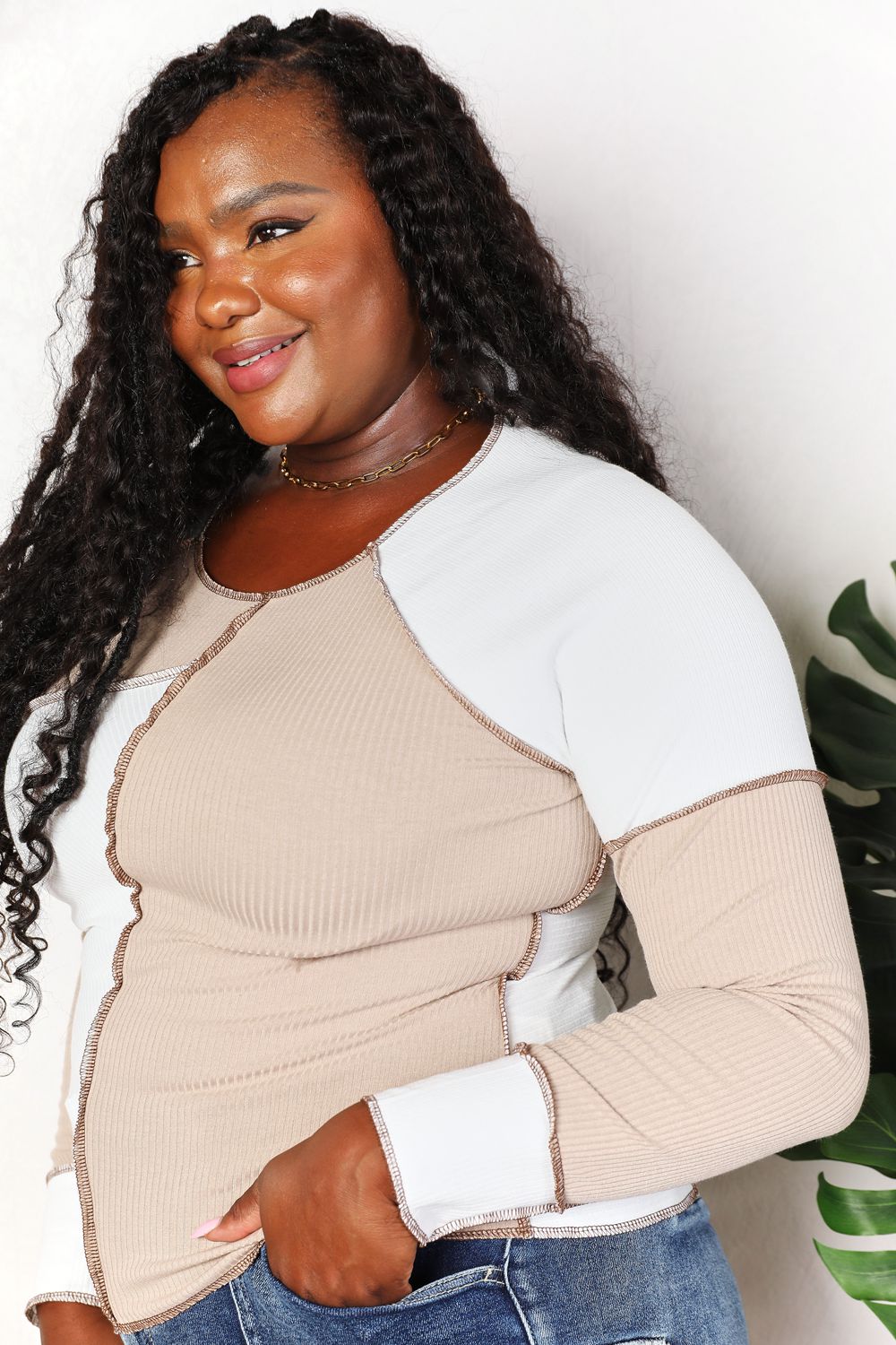 A woman with long brown hair is wearing the "Double Take Color Block Exposed Seam Top" in beige and white, paired with jeans. She stands with one hand in her pocket and the other touching her hair, smiling. A plant is visible in the background.