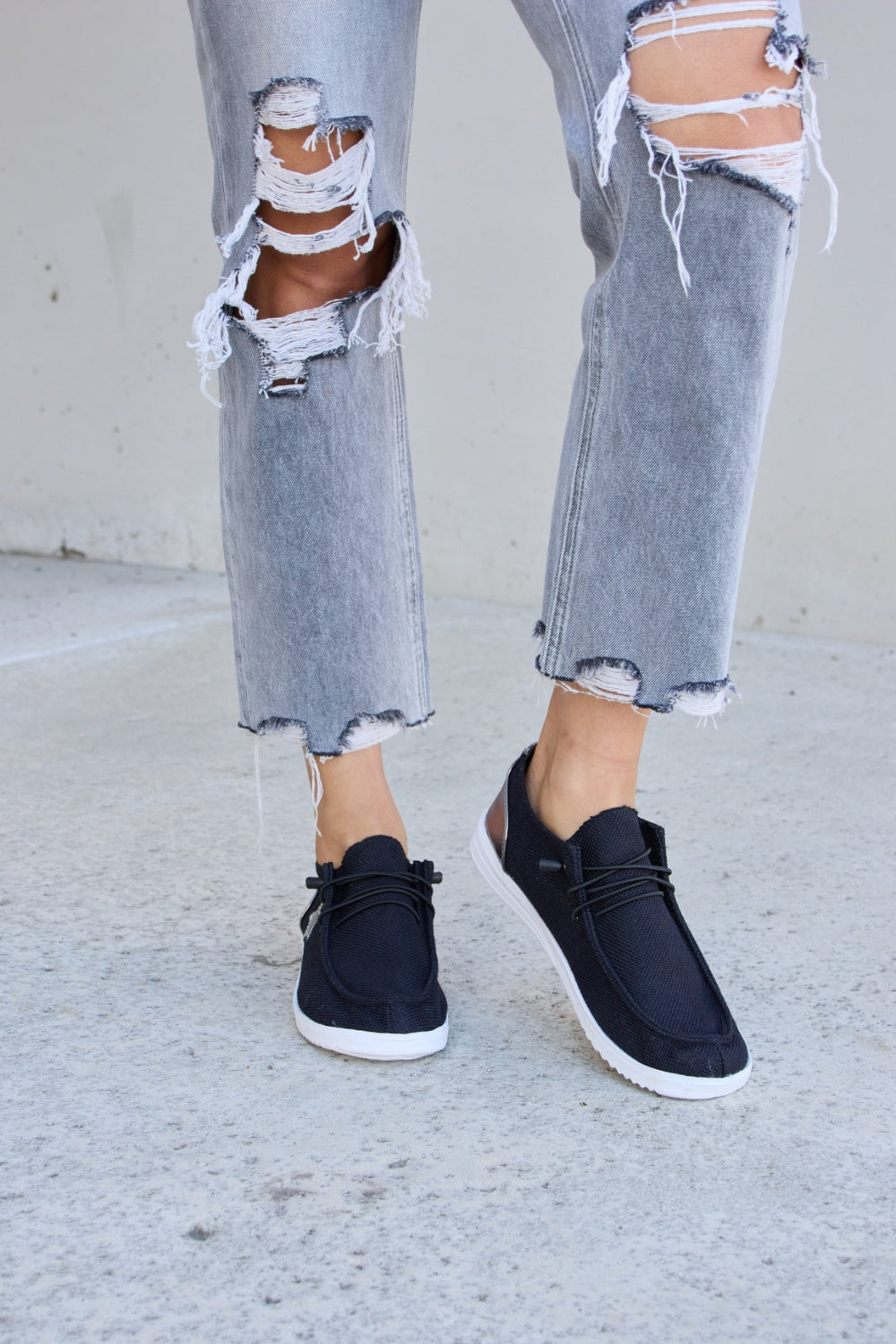 A person stands on a concrete surface, wearing distressed grey jeans and stylish Forever Link Star Lace-Up Flat Sneakers featuring black uppers and white soles.