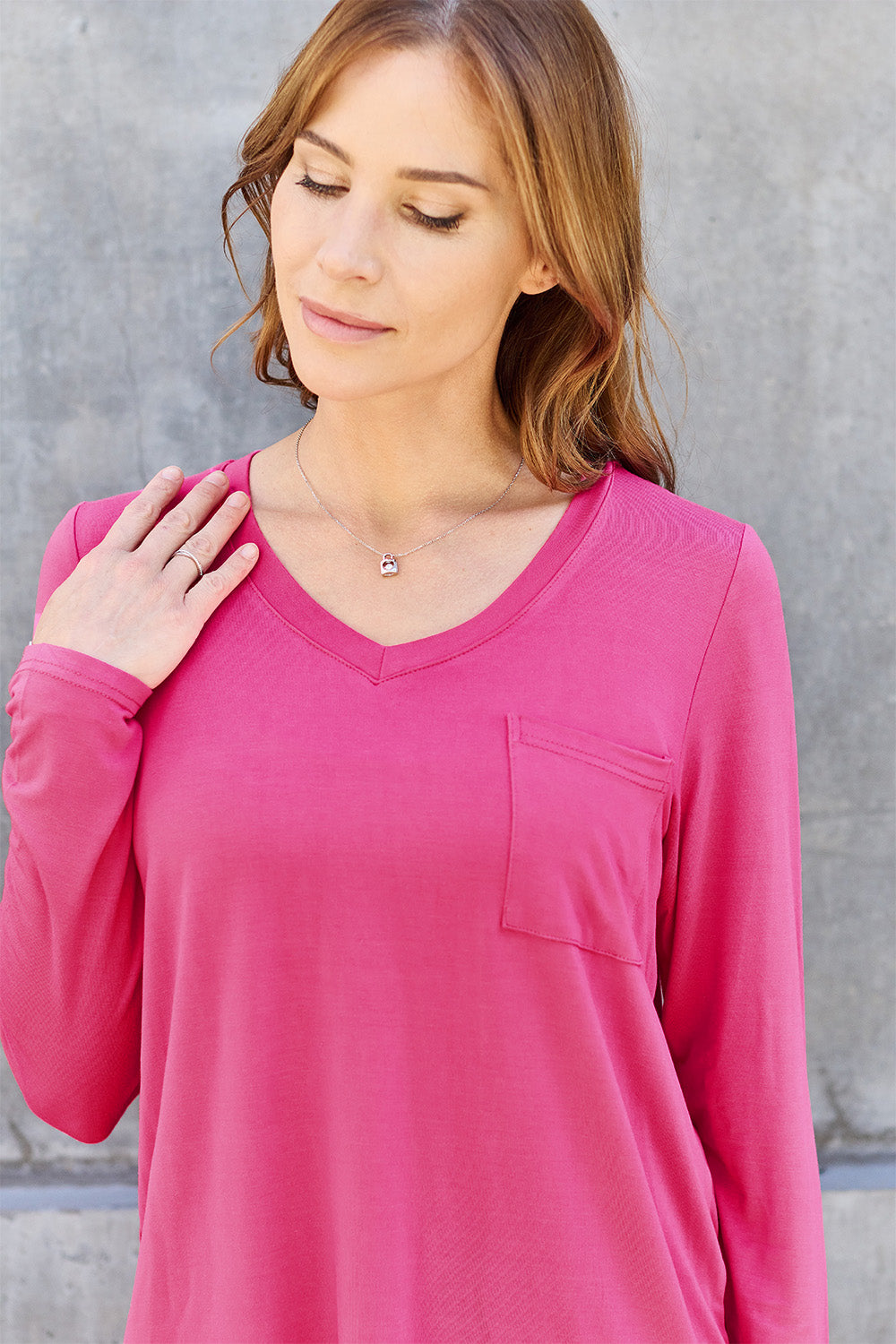 A woman with light brown hair stands against a concrete wall, wearing the Basic Bae Full Size V-Neck Long Sleeve Top in olive green and blue jeans, with a white backpack on her shoulder. Her outfit exudes basic style and is made from polyester, ensuring it’s both comfortable and suitable for machine wash cold. She is looking at the camera.