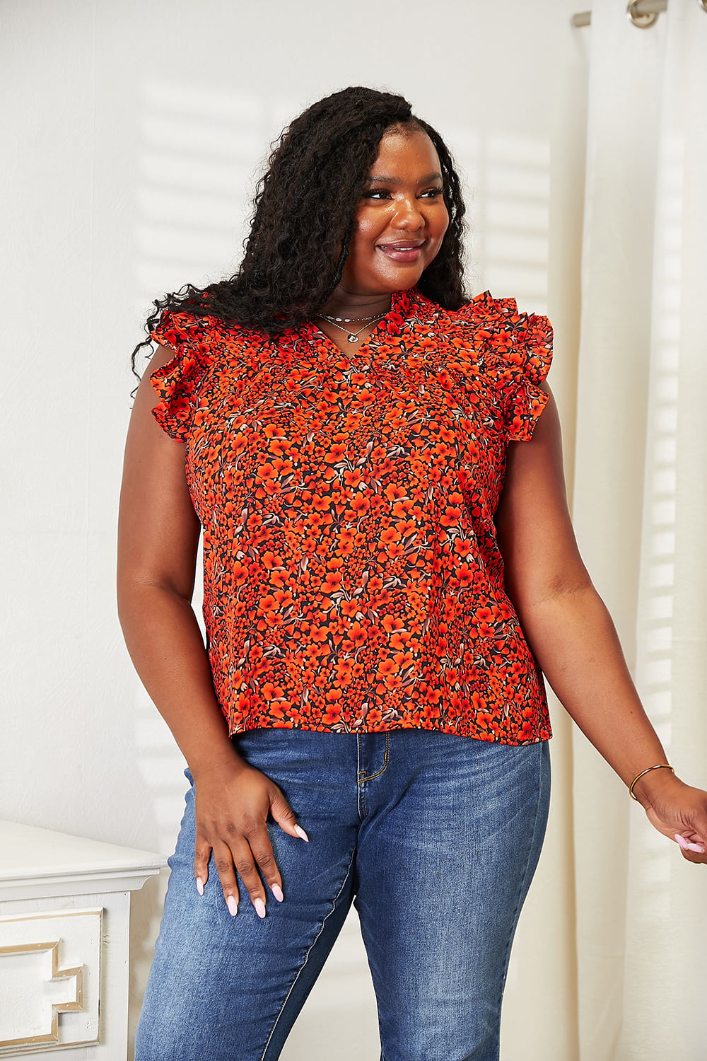 A woman wearing a chic Double Take Floral Flutter Sleeve Notched Neck Blouse and blue jeans stands smiling in a well-lit room with white walls and a curtain.