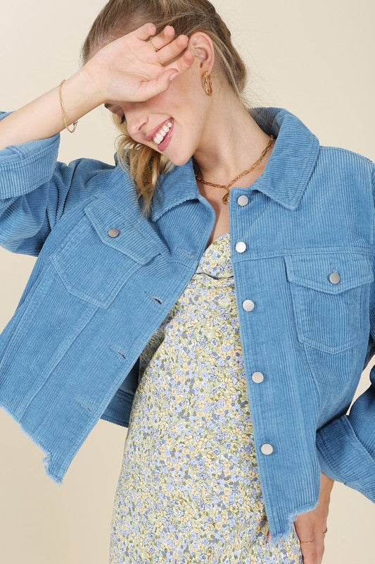 A person with long hair wears a blue frayed corduroy jacket over a floral dress, seated on a light-colored background.