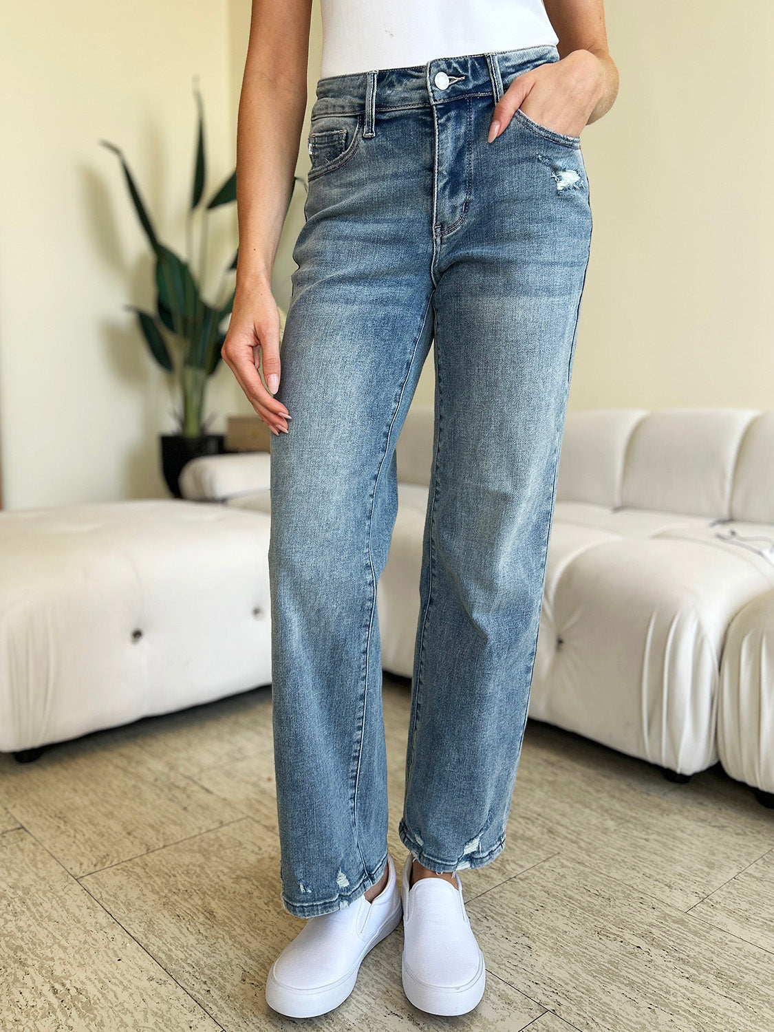 A person wearing Judy Blue Full Size Mid Rise Distressed Straight Jeans in light blue with a white top and white slip-on shoes stands in a room with a white couch and a plant.