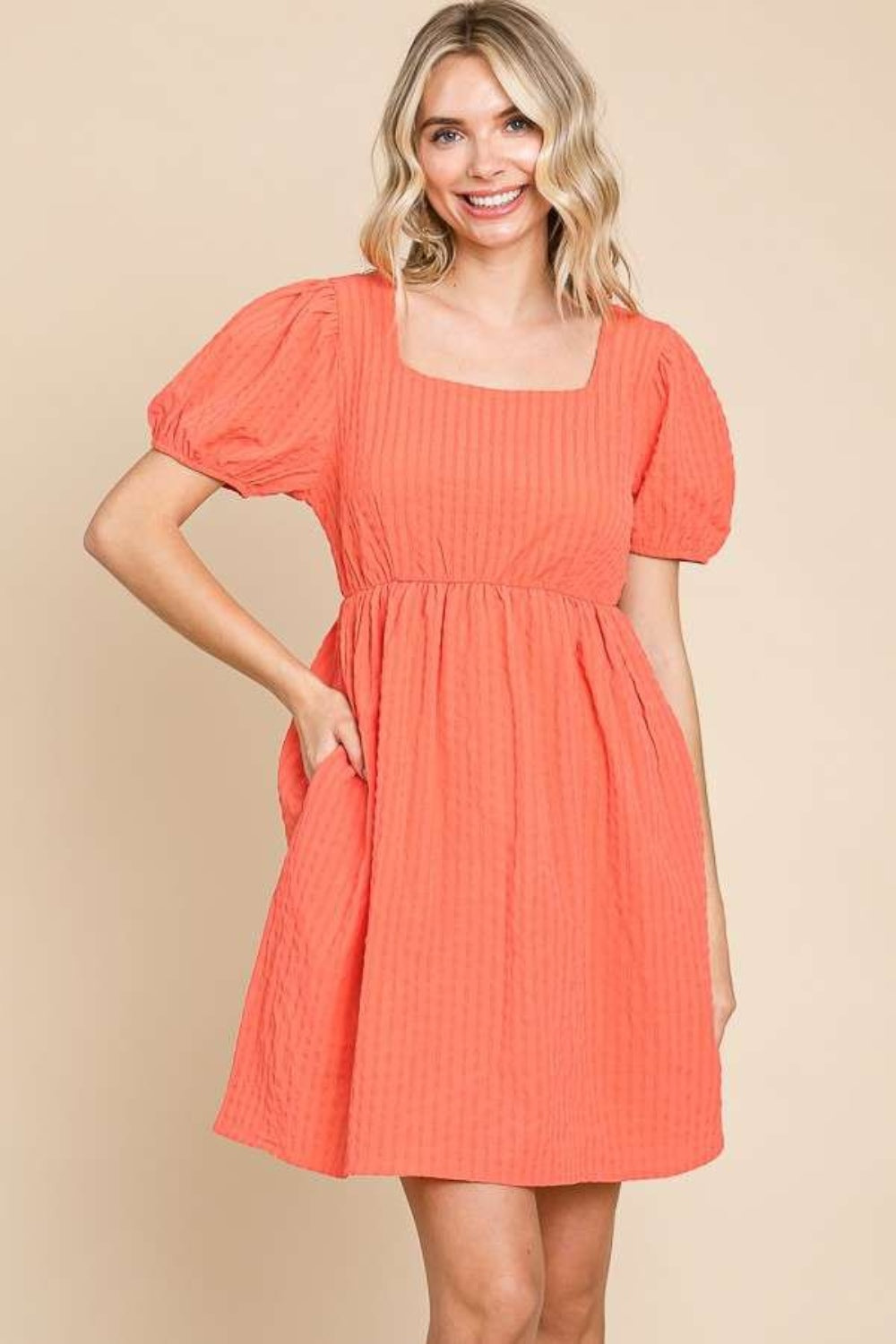 A woman is wearing a Culture Code Textured Square Neck Short Sleeve Dress: it's a stylish, short-sleeved, square-necked, orange dress with puffed sleeves. She is smiling and has one hand in her pocket.