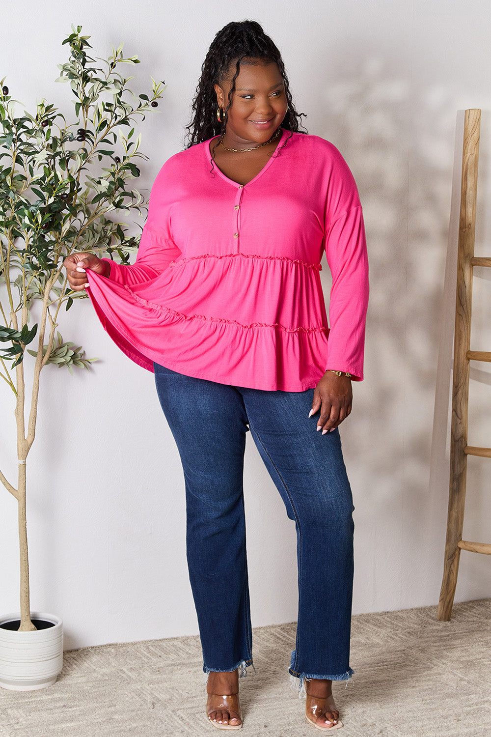 Person stands indoors near a plant, wearing the Double Take Half Button Long Sleeve Ruffle Hem Blouse in gray and blue jeans, smiling while looking to the side. The slightly stretchy fabric of the blouse adds comfort while its buttoned design offers a touch of elegance.