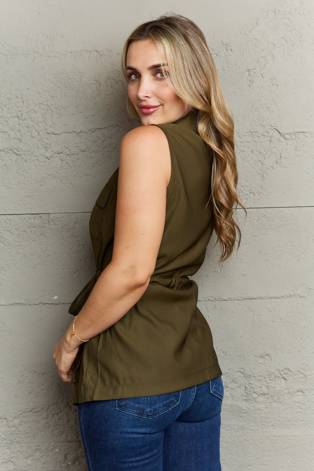 A woman with long, wavy hair wearing the Ninexis Follow The Light Sleeveless Collared Button Down Top in olive-green high-quality fabric and a belted waist stands against a textured gray wall, smiling.