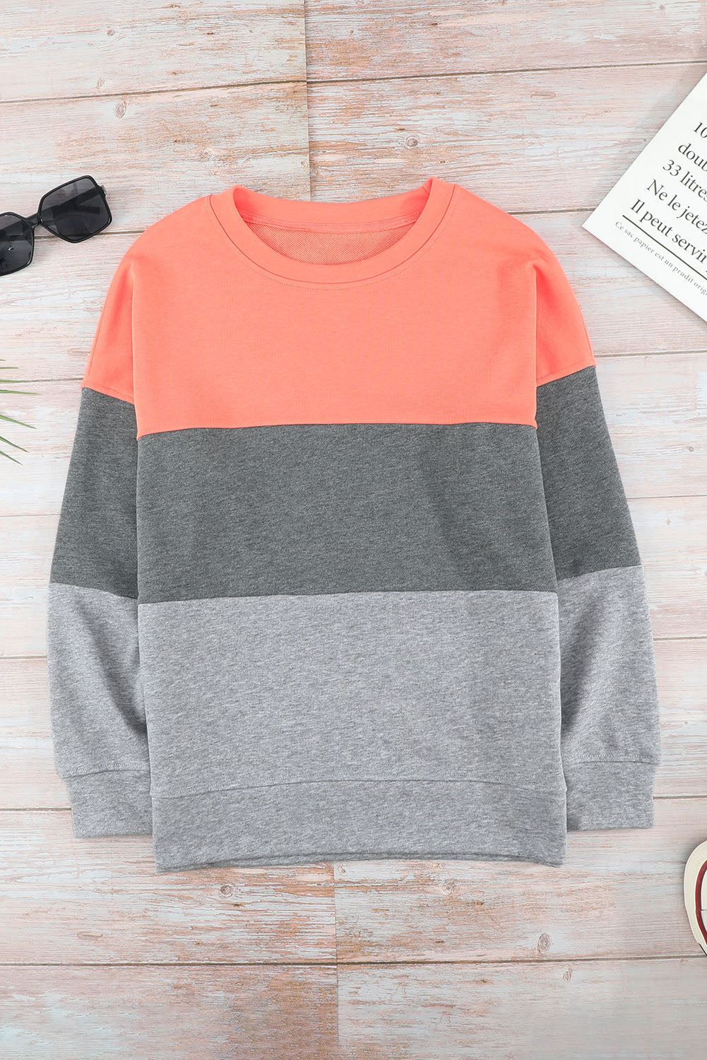 A Colorblock Gray Contrast Stitching Sweatshirt with Slits, made of comfy fabric and featuring coral, gray, and light gray sections, is placed on a wooden surface near sunglasses, a book, and some plant leaves.