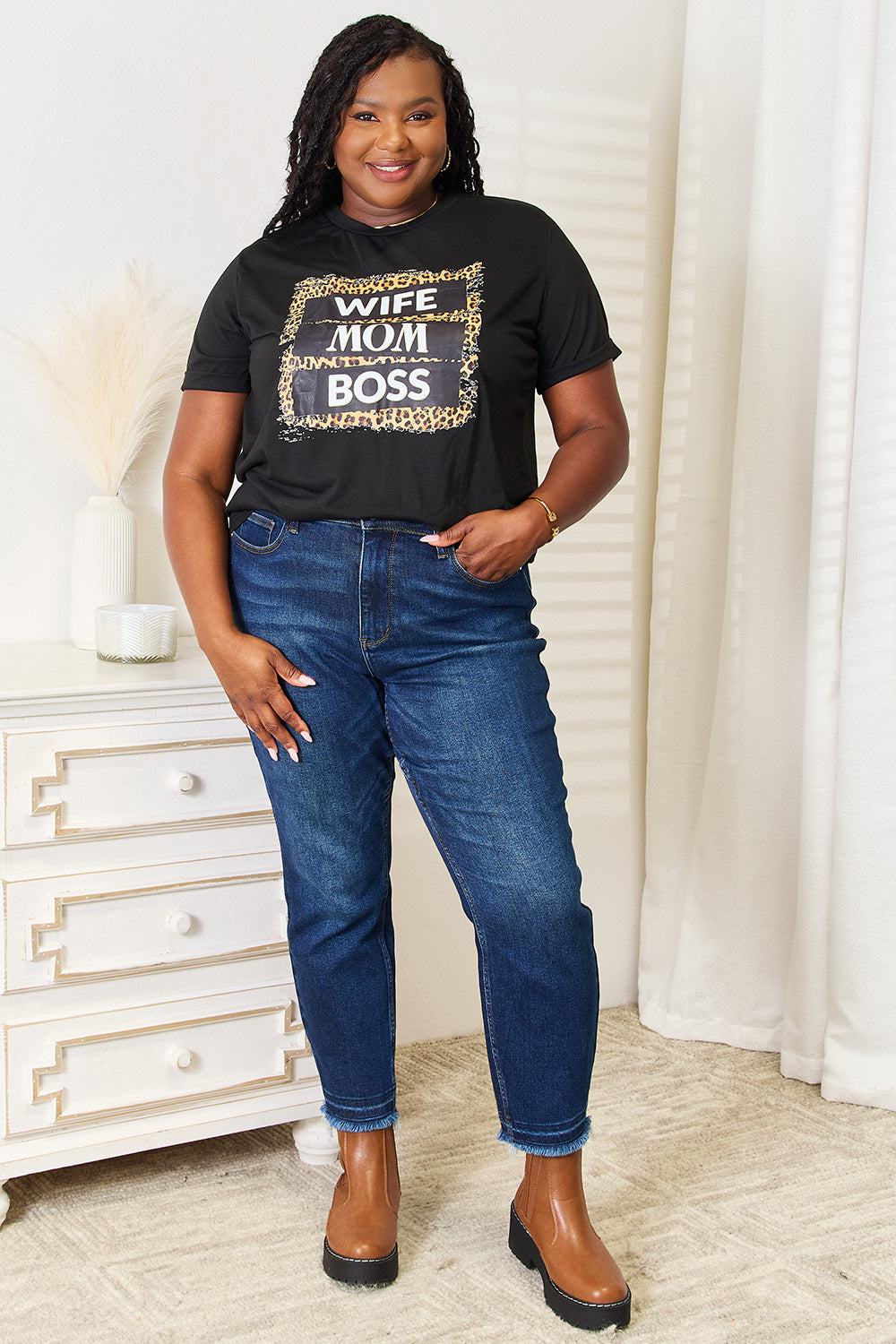 A woman beams with confidence as she dons a Simply Love WIFE MOM BOSS Leopard Graphic T-Shirt, the bold wording displayed prominently across the front. Beside a white dresser, she exudes empowerment and grace, embodying strength in her daily life.