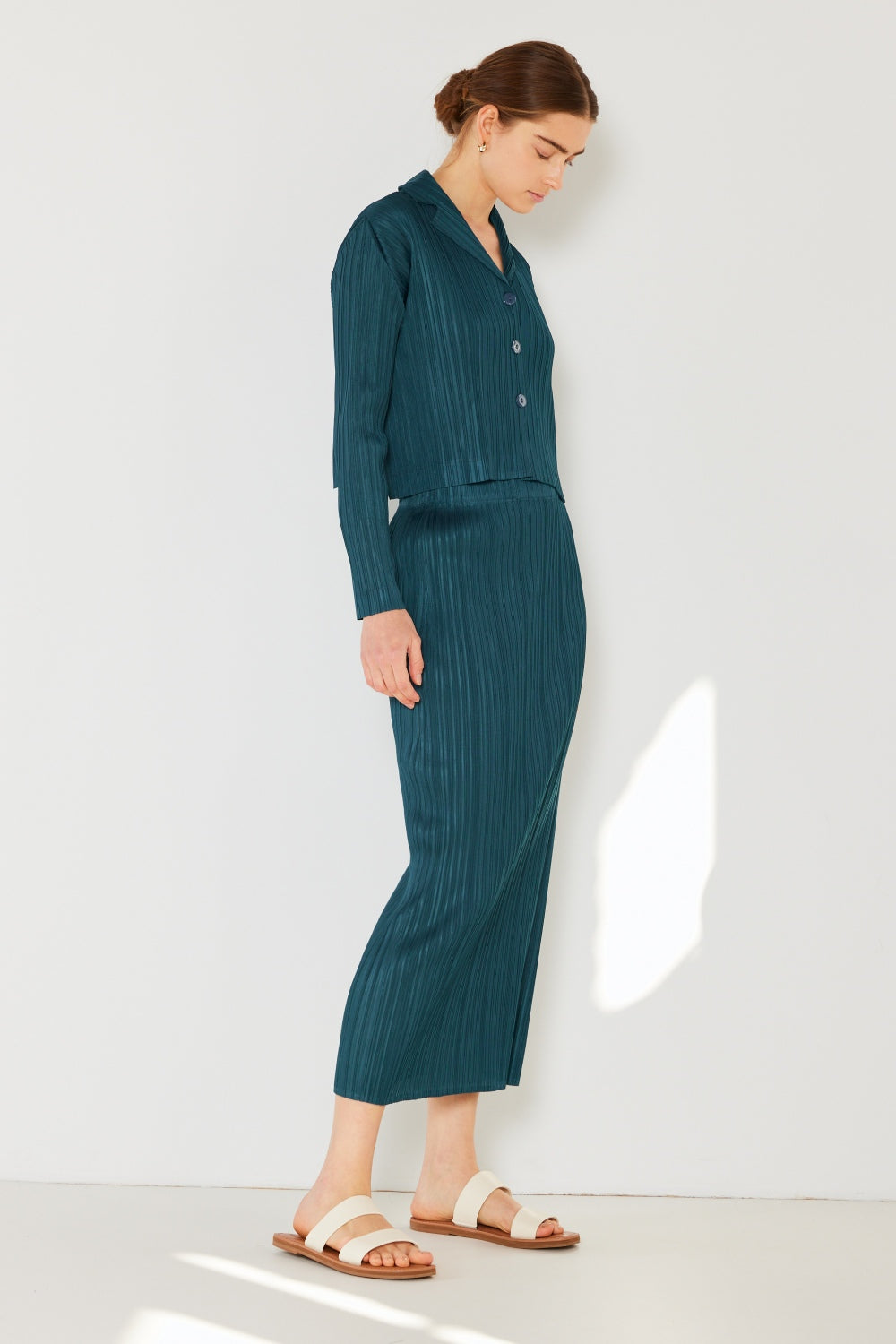 A person wearing the Marina West Swim Pleated Midi Pencil Skirt in teal, paired with a long-sleeve top, stands against a white wall, embodying a classic polished look.