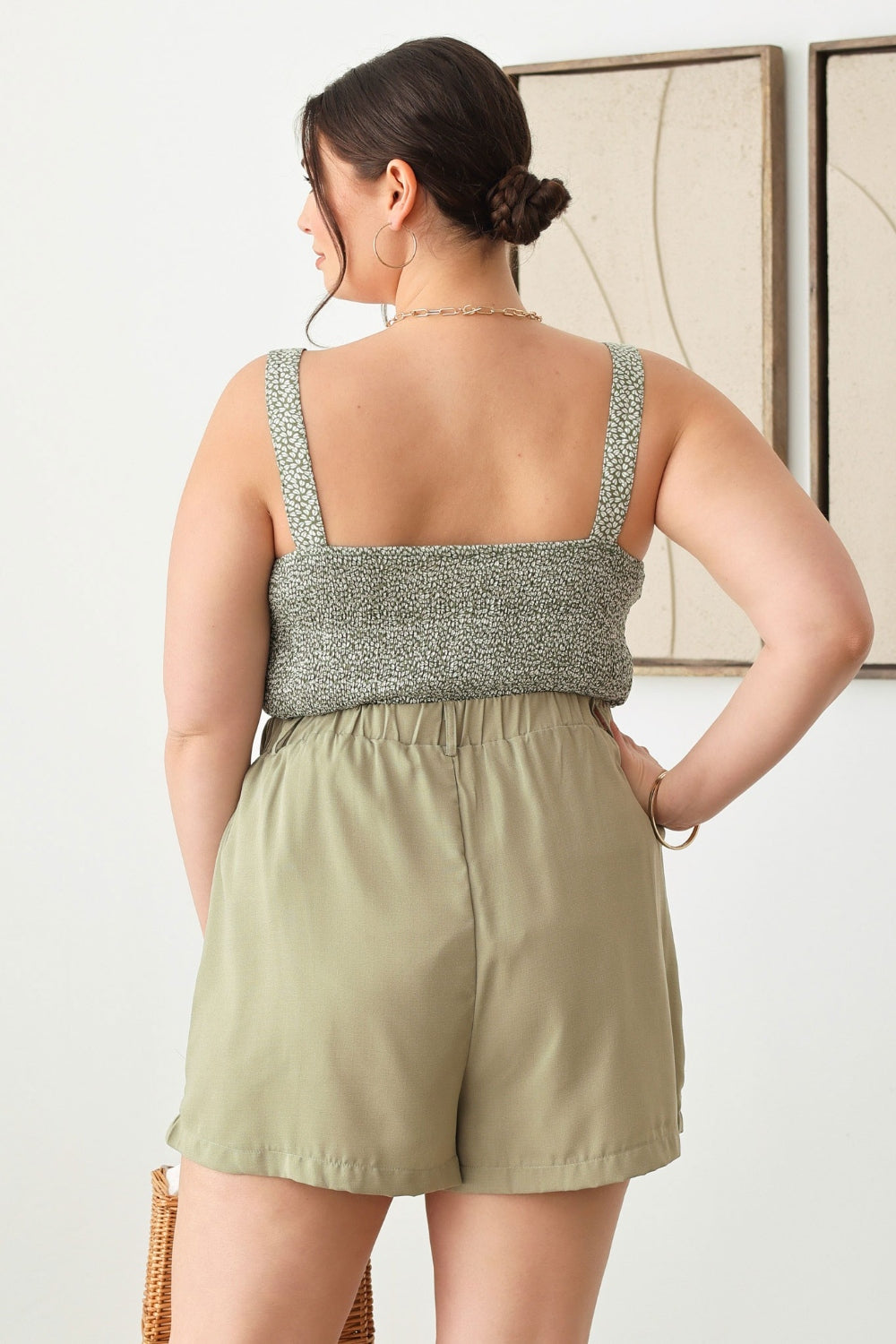 A woman in a green sleeveless top and the Zenobia Plus Size Half Elastic Waist Shorts, featuring convenient pockets, holds a woven handbag while standing indoors.