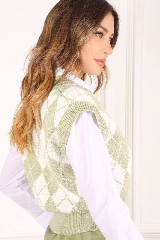 A person wearing a knitted argyle sweater vest in light green over a white shirt stands against a neutral background.