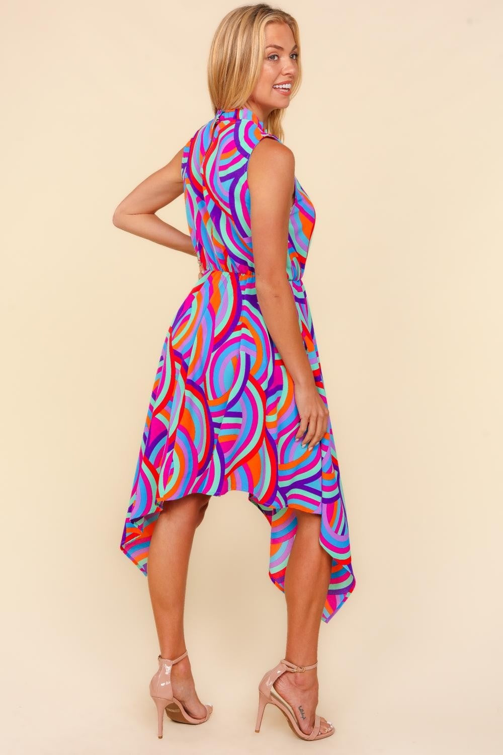 Against a beige backdrop, an individual models the Haptics Full Size Mock Neck Sleeveless Printed Dress, which features an eye-catching asymmetrical pattern. The lively blend of pink, blue, orange, and purple swirls crafts a captivating visual symphony.