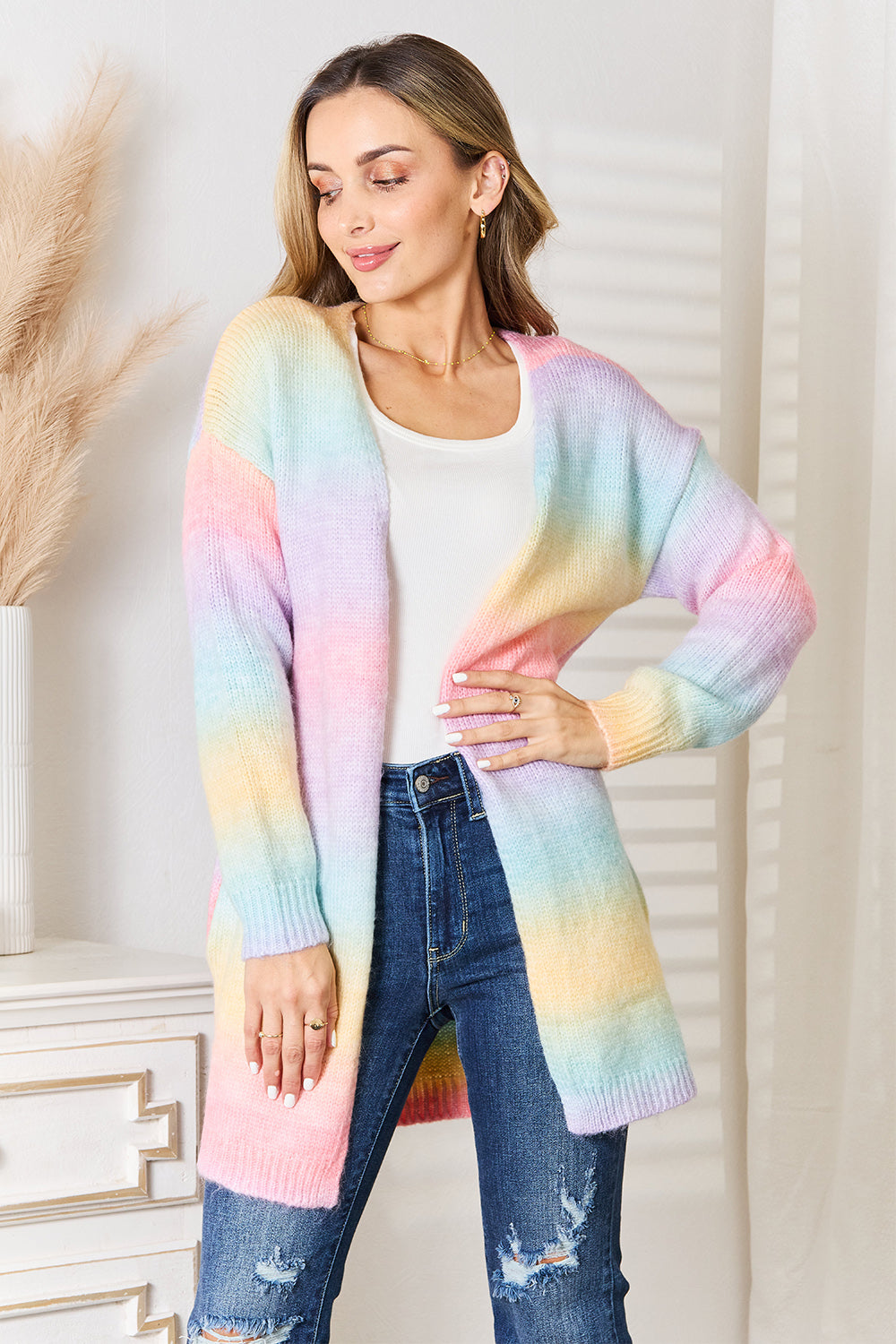 A person wearing an Angel Wings Woven Right Multicolored Gradient Open Front Longline Cardigan over a white top and blue jeans stands by a white dresser with pampas grass.