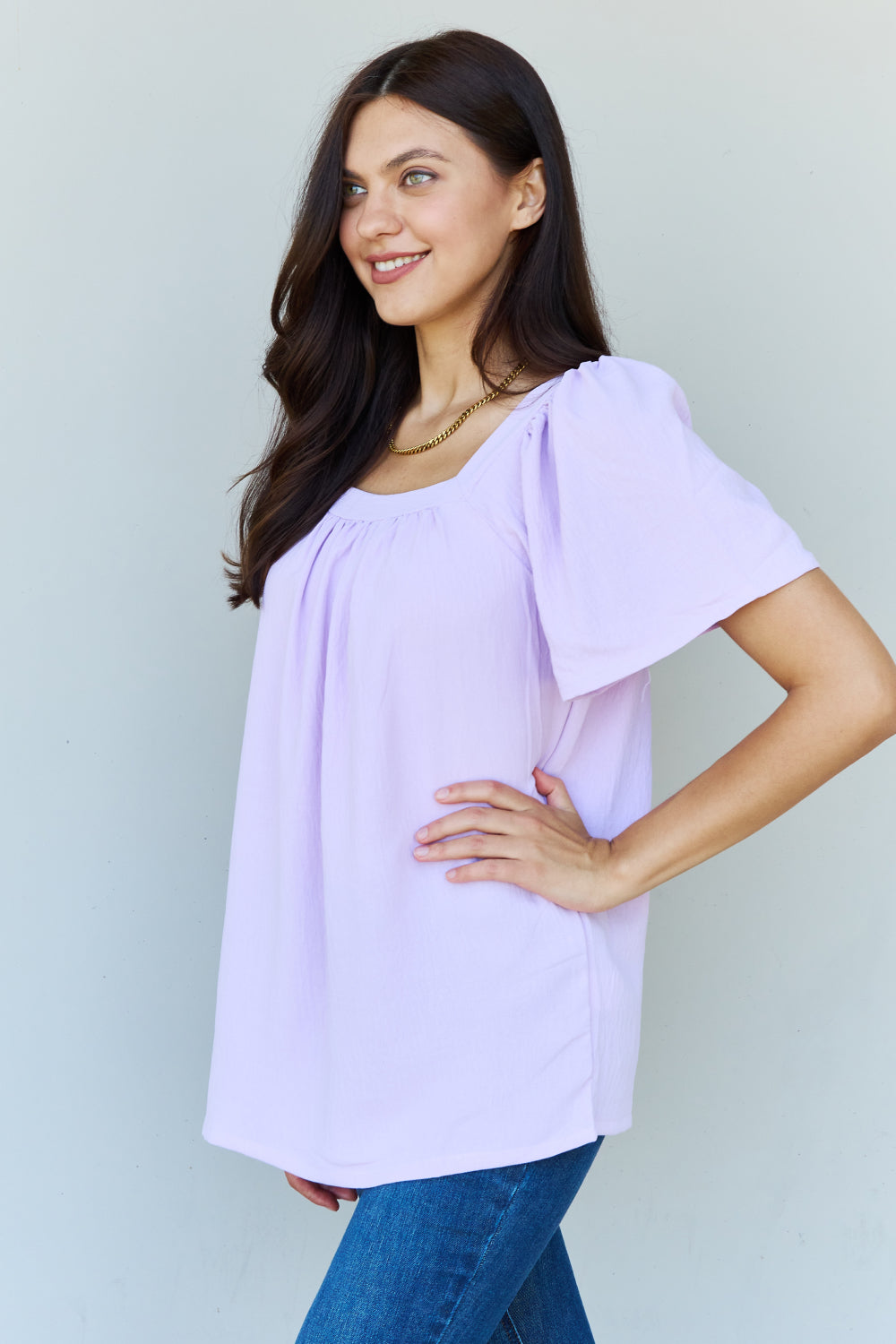 A person stands smiling in a lavender Ninexis Keep Me Close Square Neck Short Sleeve Blouse, paired with blue jeans against a plain background.