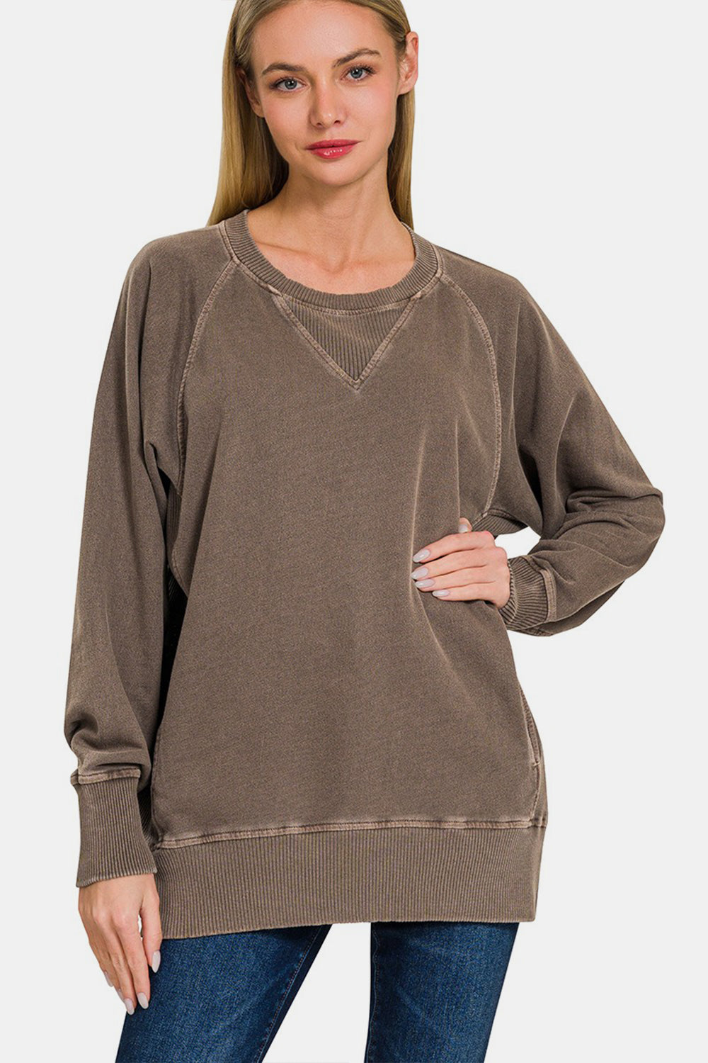 A woman wearing the Zenana Round Neck Long Sleeve Sweatshirt in beige and blue jeans stands with one hand on her hip against a neutral background.