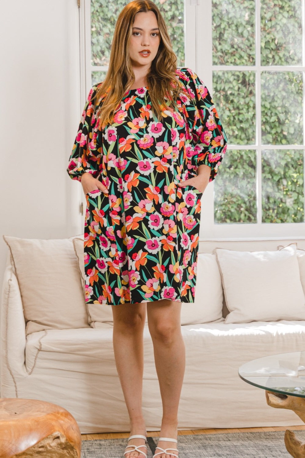 A woman wearing the stylish ODDI Full Size Floral Puff Sleeve Mini Dress stands indoors, framed by a large window with lush greenery outside.