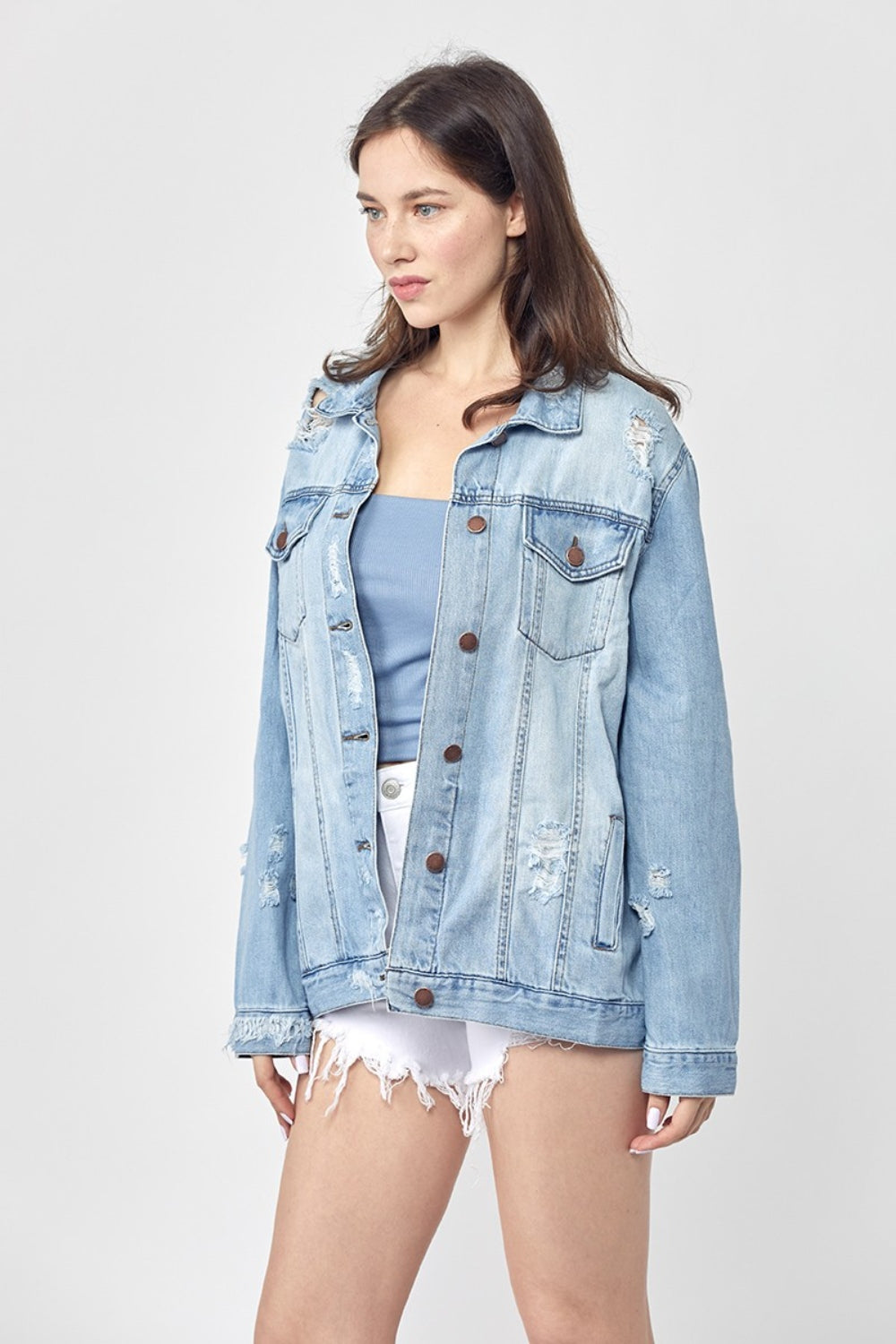 Dressed in the RISEN Full Size Distressed Long Sleeve Denim Jacket, a perfect wardrobe staple, this person effortlessly poses against a plain background while pairing it with a blue top and white shorts.