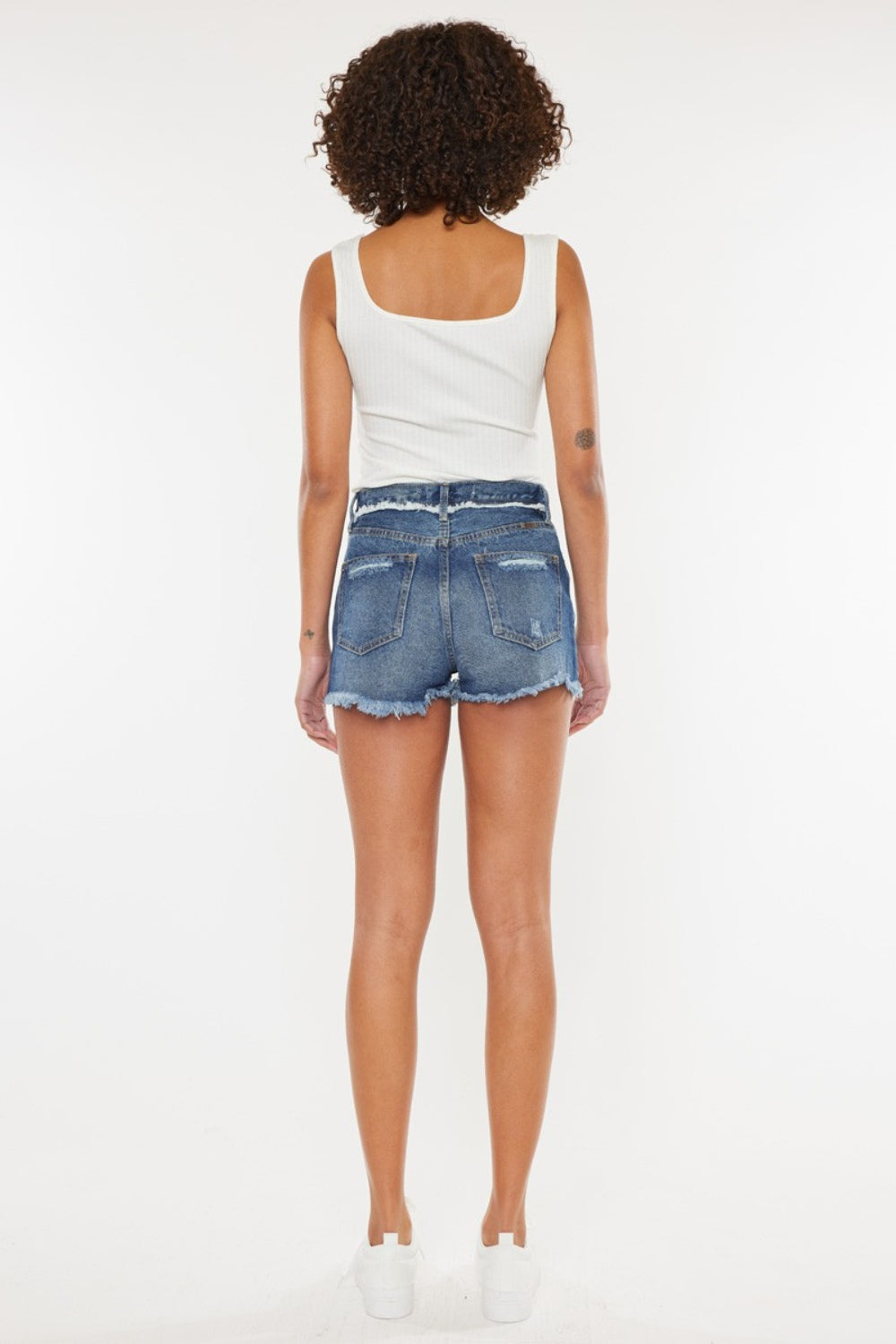 A woman with curly hair wearing a white sleeveless top and Kancan Distressed Button Fly Denim Shorts stands against a plain background, perfect for a stylish summer wardrobe.